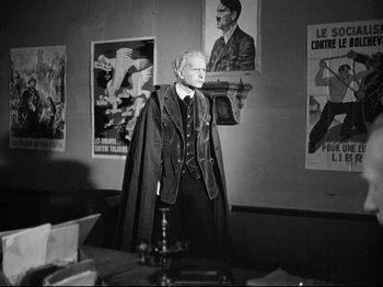 Movie still from “The Silence of the Sea” (1949), directed by Jean-Pierre Melville – An old man standing in front of a wall of posters; Medium shot, Over the shoulder angle