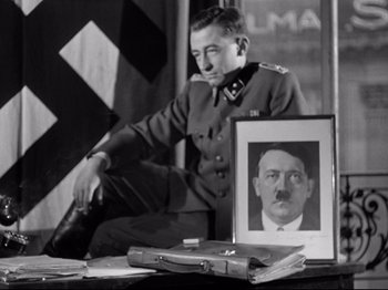 Movie still from “The Silence of the Sea” (1949), directed by Jean-Pierre Melville – A man in uniform sitting in front of a picture of adolf hitler; Medium shot, Low angle