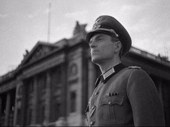 Movie still from “The Silence of the Sea” (1949), directed by Jean-Pierre Melville – A man in a military uniform standing in front of a building; Medium shot, Low angle
