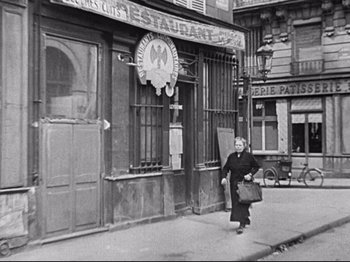 Movie still from “The Silence of the Sea” (1949), directed by Jean-Pierre Melville – An older woman walking down the street in front of a restaurant; Wide shot, High angle