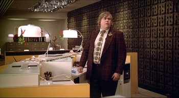 Movie still from “The Silent Partner” (1978), directed by Daryl Duke – A man in a suit and tie standing next to a desk; Medium shot, Low angle