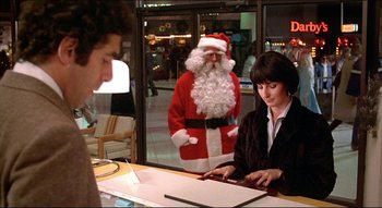 Movie still from “The Silent Partner” (1978), directed by Daryl Duke – A man dressed as santa claus stands next to a woman as she looks at a tablet; Medium shot, Over the shoulder angle