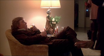 Movie still from “The Silent Partner” (1978), directed by Daryl Duke – A man sitting on top of a couch drinking a glass of wine; Medium shot, Over the shoulder angle