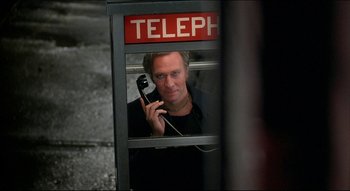 Movie still from “The Silent Partner” (1978), directed by Daryl Duke – A man holding a phone up to his ear while sitting in a phone booth; Close Up shot, Overhead angle