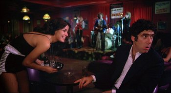 Movie still from “The Silent Partner” (1978), directed by Daryl Duke – A man and a woman sitting at a table in front of a crowd; Medium shot, Over the shoulder angle