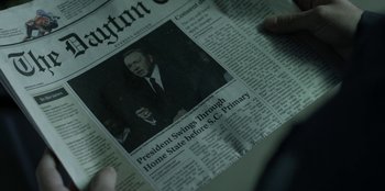 Movie still from “House of Cards” (2013), created by Beau Willimon – A person holding a newspaper with a picture of a man in a black suit; Extreme Close Up shot, Overhead angle