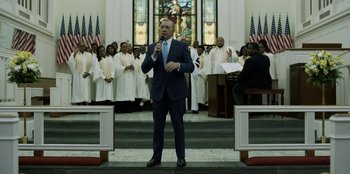 Movie still from “House of Cards” (2013), created by Beau Willimon – A man in a blue suit is speaking in front of a group of people; Medium shot, Low angle