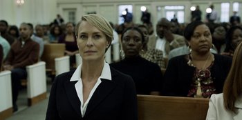Movie still from “House of Cards” (2013), created by Beau Willimon – A woman in a black suit sitting in front of a group of people; Medium shot, Low angle