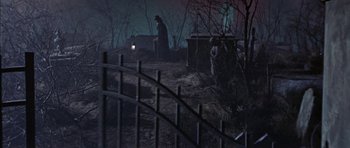 Movie still from “The Skull” (1965), directed by Freddie Francis – A man in a black coat standing next to a fence; Wide shot, High angle