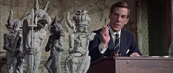 Movie still from “The Skull” (1965), directed by Freddie Francis – A man in a suit and tie speaking at a podium in front of statues; Medium shot, Low angle