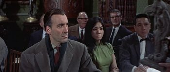 Movie still from “The Skull” (1965), directed by Freddie Francis – A man and a woman are standing next to each other in front of other men; Medium shot, Low angle