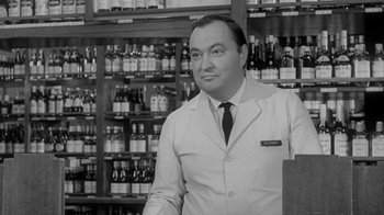 Movie still from “The Slender Thread” (1965), directed by Sydney Pollack – A man standing in front of a store filled with bottles of alcohol; Medium shot, Low angle