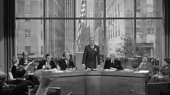 Movie still from “The Solid Gold Cadillac” (1956), directed by Richard Quine – A man standing in front of a group of people in a room; Wide shot, Over the shoulder angle