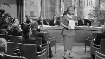 Movie still from “The Solid Gold Cadillac” (1956), directed by Richard Quine – A woman is holding papers while standing in front of a group of people; Wide shot, Over the shoulder angle