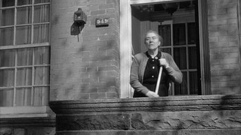 Movie still from “The Solid Gold Cadillac” (1956), directed by Richard Quine – An older woman standing on the porch of a brick building; Medium shot, Low angle