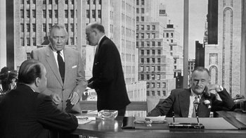 Movie still from “The Solid Gold Cadillac” (1956), directed by Richard Quine – A group of men standing around a table in front of a window; Medium shot, Over the shoulder angle