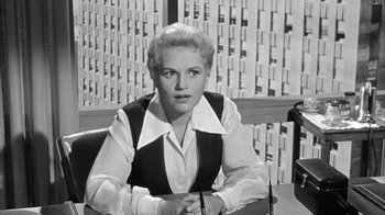 Movie still from “The Solid Gold Cadillac” (1956), directed by Richard Quine – A woman sitting at a desk in front of papers; Medium shot, Low angle
