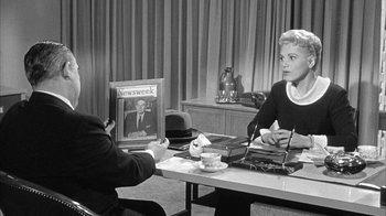 Movie still from “The Solid Gold Cadillac” (1956), directed by Richard Quine – A woman sitting at a table holding a magazine; Medium shot, Over the shoulder angle