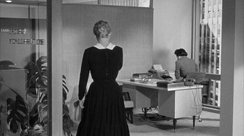 Movie still from “The Solid Gold Cadillac” (1956), directed by Richard Quine – A woman in a black dress standing in front of a desk; Medium shot, Over the shoulder angle