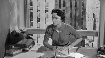Movie still from “The Solid Gold Cadillac” (1956), directed by Richard Quine – An old black and white photo of a woman at a desk; Medium shot, Low angle