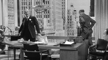 Movie still from “The Solid Gold Cadillac” (1956), directed by Richard Quine – A man in a suit and tie standing in front of a desk; Wide shot, Over the shoulder angle