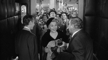 Movie still from “The Solid Gold Cadillac” (1956), directed by Richard Quine – An old black and white photo of a crowd of men and a woman; Medium shot, Over the shoulder angle