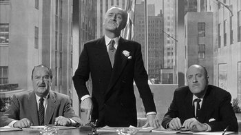 Movie still from “The Solid Gold Cadillac” (1956), directed by Richard Quine – An old photo of a man standing at a table in front of two other men; Medium shot, Low angle