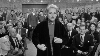 Movie still from “The Solid Gold Cadillac” (1956), directed by Richard Quine – An old photo of a woman standing in front of a crowd; Medium shot, Over the shoulder angle