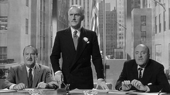 Movie still from “The Solid Gold Cadillac” (1956), directed by Richard Quine – A black and white photo of a man standing at a conference table; Medium shot, Over the shoulder angle