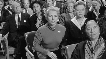 Movie still from “The Solid Gold Cadillac” (1956), directed by Richard Quine – A woman sitting in a crowd of people; Medium shot, Over the shoulder angle