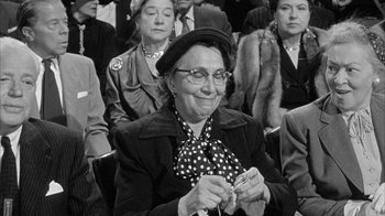 Movie still from “The Solid Gold Cadillac” (1956), directed by Richard Quine – An older woman sitting in front of a group of people; Medium shot, Over the shoulder angle
