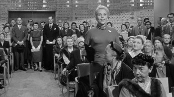 Movie still from “The Solid Gold Cadillac” (1956), directed by Richard Quine – An older woman standing in front of a group of people; Medium shot, Over the shoulder angle