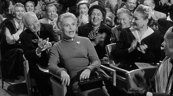 Movie still from “The Solid Gold Cadillac” (1956), directed by Richard Quine – An older woman sitting in front of a crowd of people; Medium shot, Over the shoulder angle
