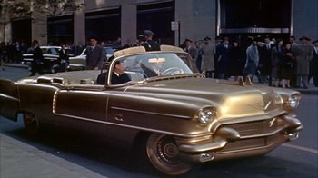 Movie still from “The Solid Gold Cadillac” (1956), directed by Richard Quine – An old car is driving down the street; Wide shot, High angle