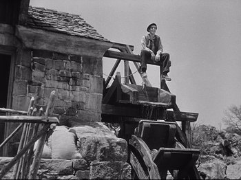 Movie still from “The Song of Bernadette” (1943), directed by Henry King – A man sitting on a wooden platform next to a building; Wide shot, Low angle