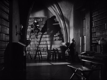 Movie still from “The Song of Bernadette” (1943), directed by Henry King – A man is sitting on a ladder in front of a book shelf; Wide shot, Low angle