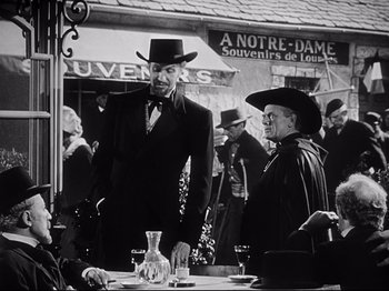 Movie still from “The Song of Bernadette” (1943), directed by Henry King – A man in a suit and hat standing next to a table; Medium shot, Low angle