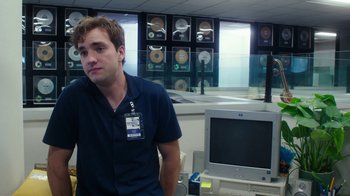 Movie still from “The Space Between” (2021), directed by Rachel Winter – A man in a blue shirt is in front of a computer; Medium shot, Over the shoulder angle