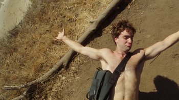 Movie still from “The Space Between” (2021), directed by Rachel Winter – A shirtless man holding a stick in the dirt; Medium shot, Overhead angle