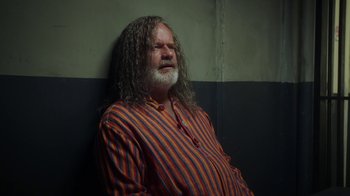 Movie still from “The Space Between” (2021), directed by Rachel Winter – A man with long hair and a beard sitting on a wall; Medium shot, Over the shoulder angle