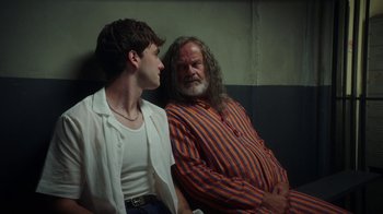 Movie still from “The Space Between” (2021), directed by Rachel Winter – A man sitting next to another man on a couch; Medium shot, Over the shoulder angle