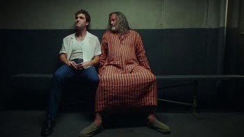 Movie still from “The Space Between” (2021), directed by Rachel Winter – Two men sitting next to each other on a bench; Medium shot, Low angle