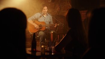 Movie still from “The Space Between” (2021), directed by Rachel Winter – A man sitting in front of a microphone playing an acoustic guitar; Medium shot, Over the shoulder angle