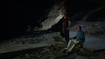 Movie still from “The Space Between” (2021), directed by Rachel Winter – Two men are sitting on the ground in a cave; Wide shot, Low angle