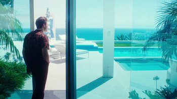 Movie still from “The Space Between” (2021), directed by Rachel Winter – A man standing in front of an open window looking out at the ocean; Wide shot, Low angle