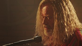 Movie still from “The Space Between” (2021), directed by Rachel Winter – A man with long hair is holding a microphone; Close Up shot, Low angle