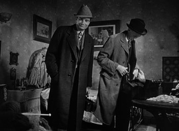 Movie still from “The Spiral Staircase” (1946), directed by Robert Siodmak – Two men in suits and hats standing next to each other in a cluttered room; Medium shot, Low angle