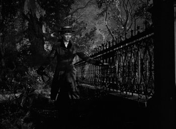 Movie still from “The Spiral Staircase” (1946), directed by Robert Siodmak – An old photo of a woman in a hat and a long coat standing in front of an iron fence; Wide shot, Low angle