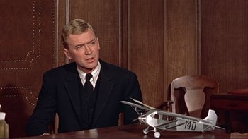 Movie still from “The Spirit of St. Louis” (1957), directed by Billy Wilder – A man in a suit and tie sitting at a table with a model airplane; Medium shot, Over the shoulder angle