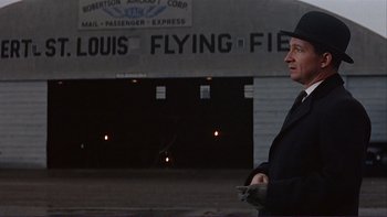 Movie still from “The Spirit of St. Louis” (1957), directed by Billy Wilder – A man in a black hat standing in front of an airplane hangar; Medium shot, Low angle